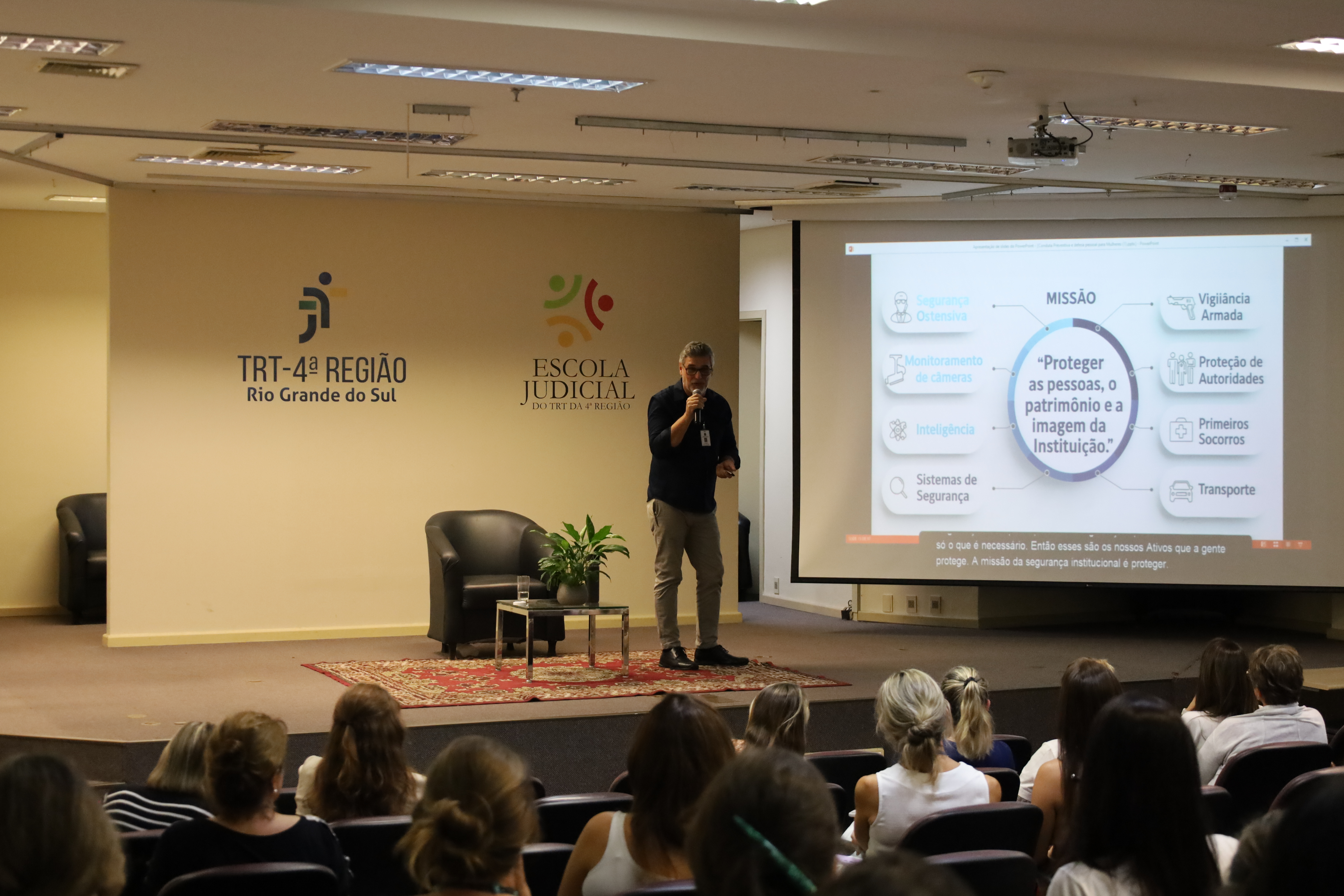 Auditório com palco onde o palestrante segura um microfone e apresenta um slide projetado em tela grande. No slide, aparece a missão da segurança institucional, com ícones e palavras-chave. Ao lado, há uma poltrona e uma pequena mesa com planta decorativa. Na parede, estão as marcas do TRT e da Escola Judicial. O público, majoritariamente feminino, está sentado em fileiras, assistindo com atenção à apresentação.