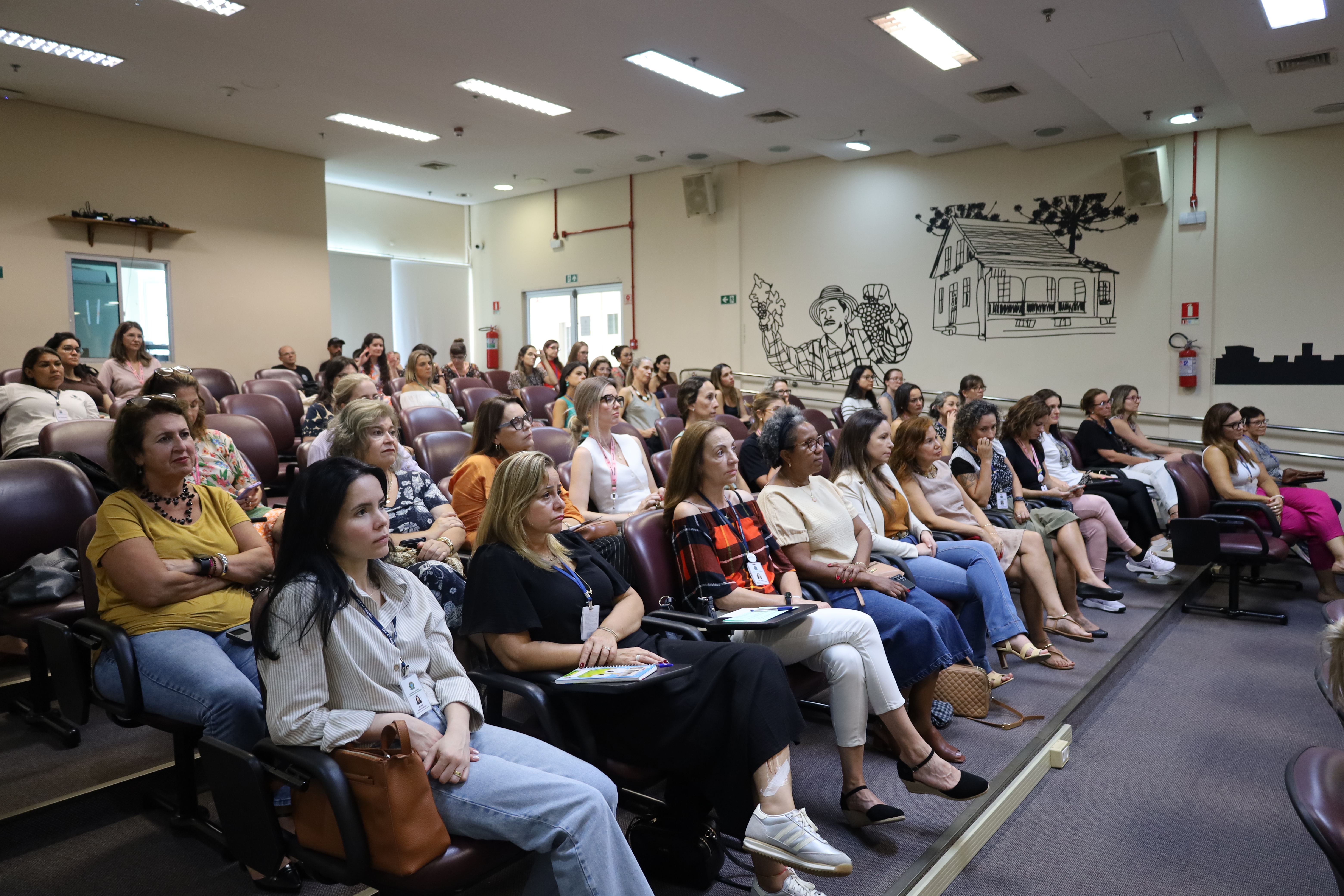 Auditório visto de lado, com várias fileiras de cadeiras ocupadas por mulheres atentas à atividade. O ambiente é bem iluminado e organizado, com paredes claras e ilustrações decorativas ao fundo. Algumas participantes seguram cadernos ou celulares.