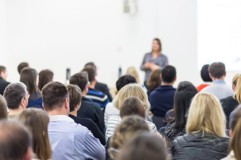 Imagem desfocada de mulher, à frente de sala de aula. Ela tem a pele branca e cabelos escuros, lisos, na altura dos ombros. Há vários estudantes sentados. Eles aparecem de costas.