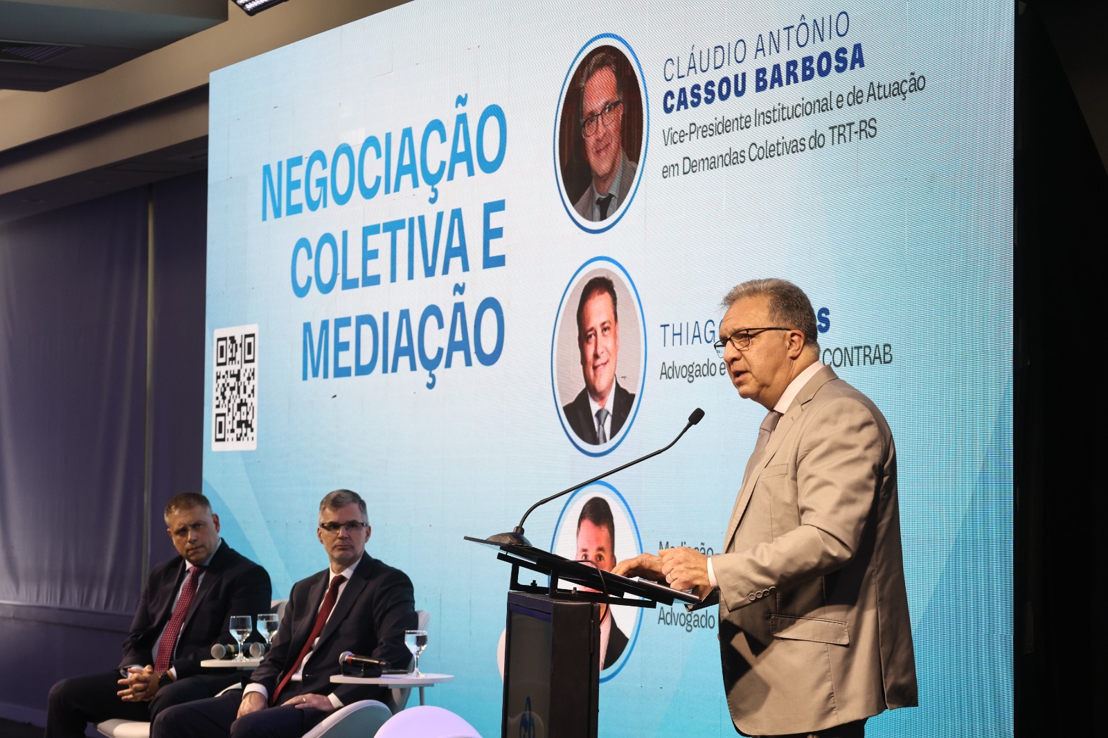 Thiago, Gustavo e Cassou no palco do evento. Ao fundo aparece um telão com os participantes e o tema do painel. Cassou está de pé, na direita da imagem, falando no púlpito.