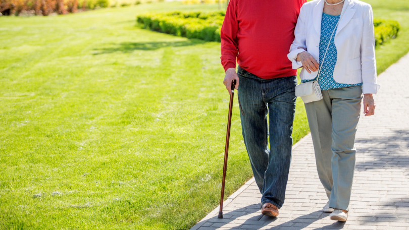 Homem e mulher passeando em parque. Ele segura uma bengala e está de braços dados com ela. Ambos têm a pele clara. Ele usa calça jeans e blusão vermelho. Ela está com calça cinza, blusa azul e casaco branco. Não há imagem dos rostos.