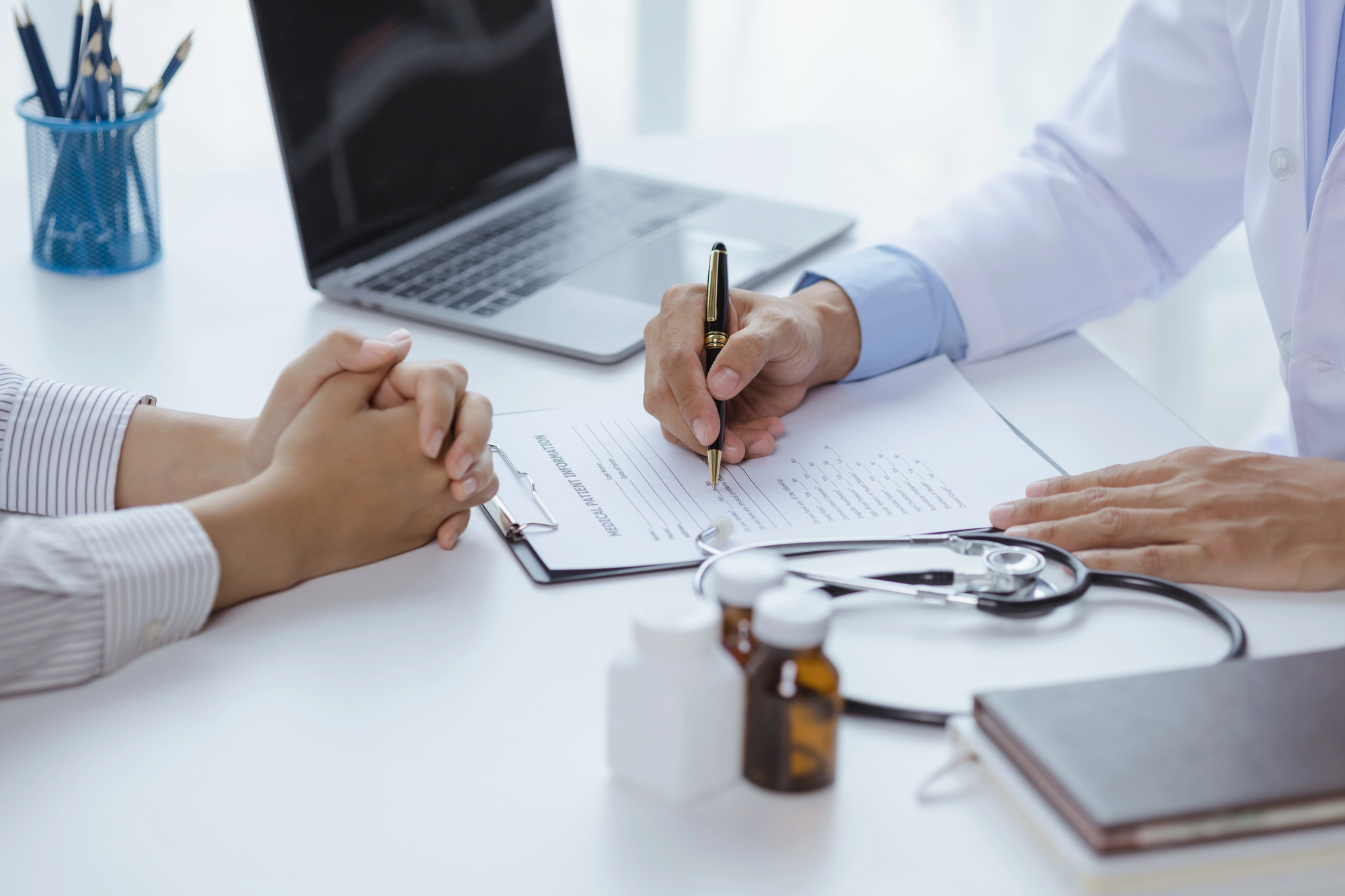 Close em médico e paciente, sentados na mesa, apenas as mãos à mostra. A imagem não mostra o rosto das pessoas. O médico preenche um documento à caneta e o paciente aguarda com as mãos unidas sobre a mesa.