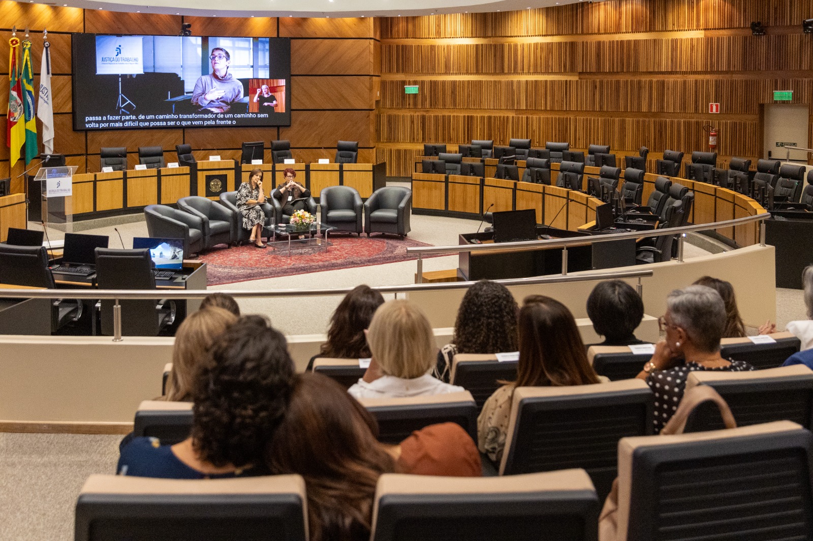 telão ao fundo exibe vídeo da entrevista com Andréa Nocchi. Público aparece de costas assistindo ao vídeo. Na nave ao centro, Beatriz e Ana Naiara estão sentadas em sofás