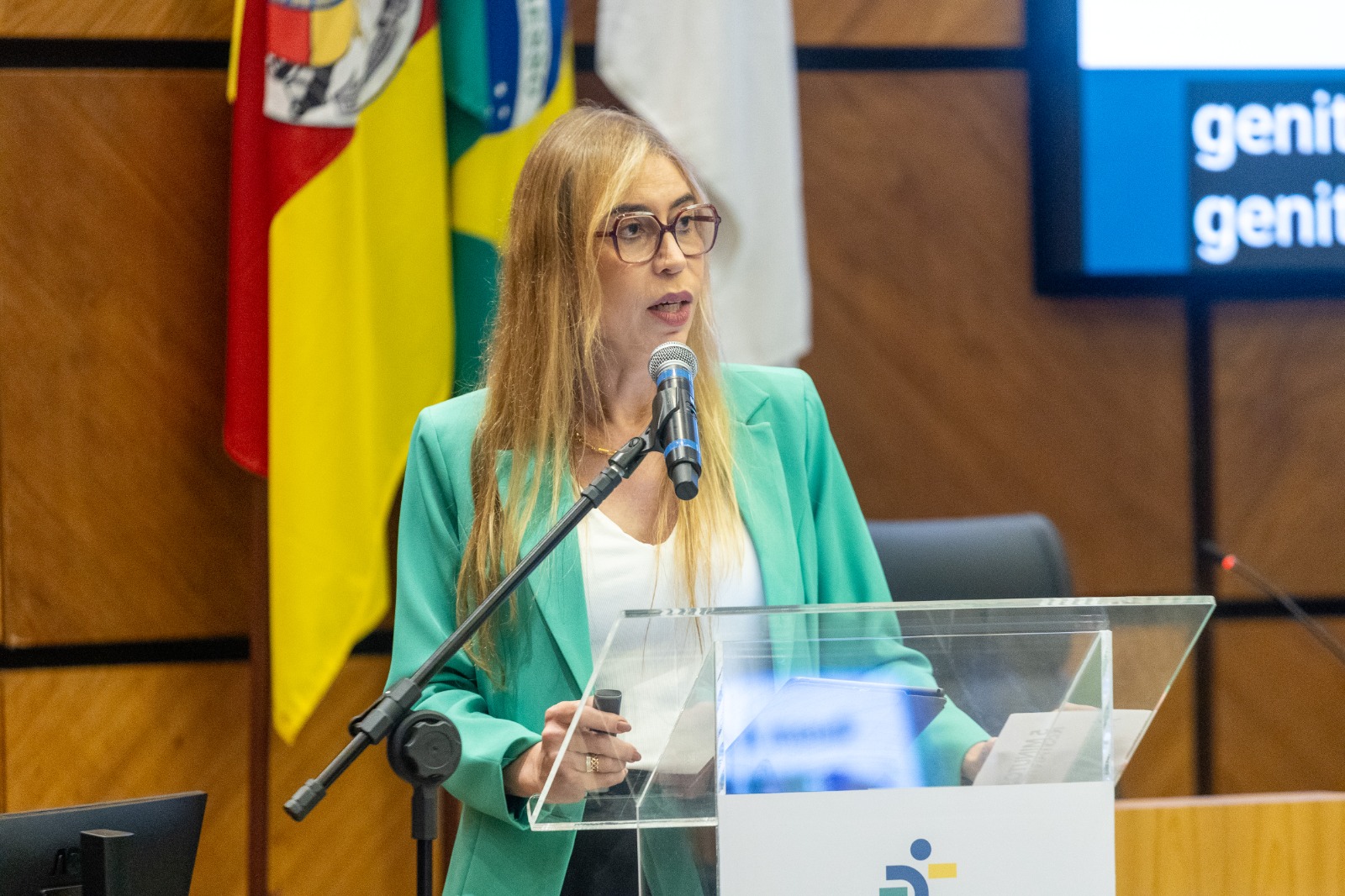 Alê é loira, branca, usa óculos de aro grande e vesta blazer verde.