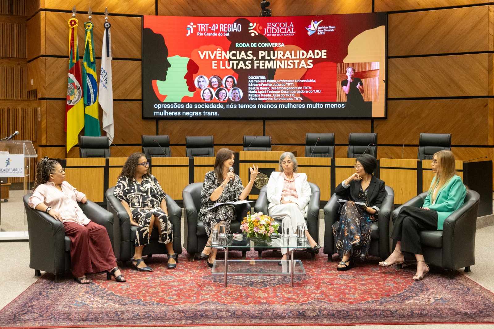 Seis mulheres sentadas em cadeiras na nave do Plenário, com a arte do evento no telão ao fundo
