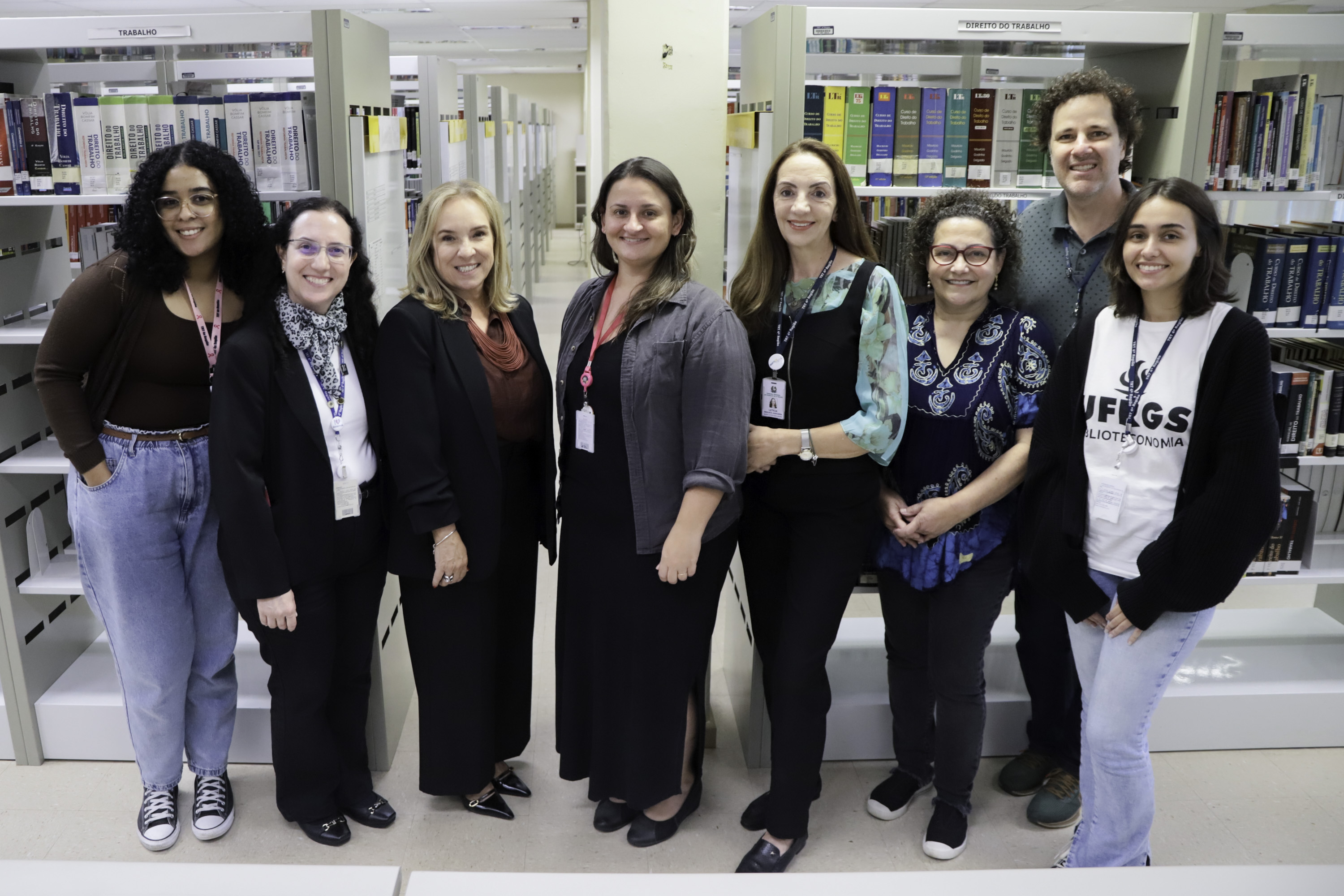 Oito pessoas de pé posam para a foto. O acervo físico da biblioteca aparece ao fundo.