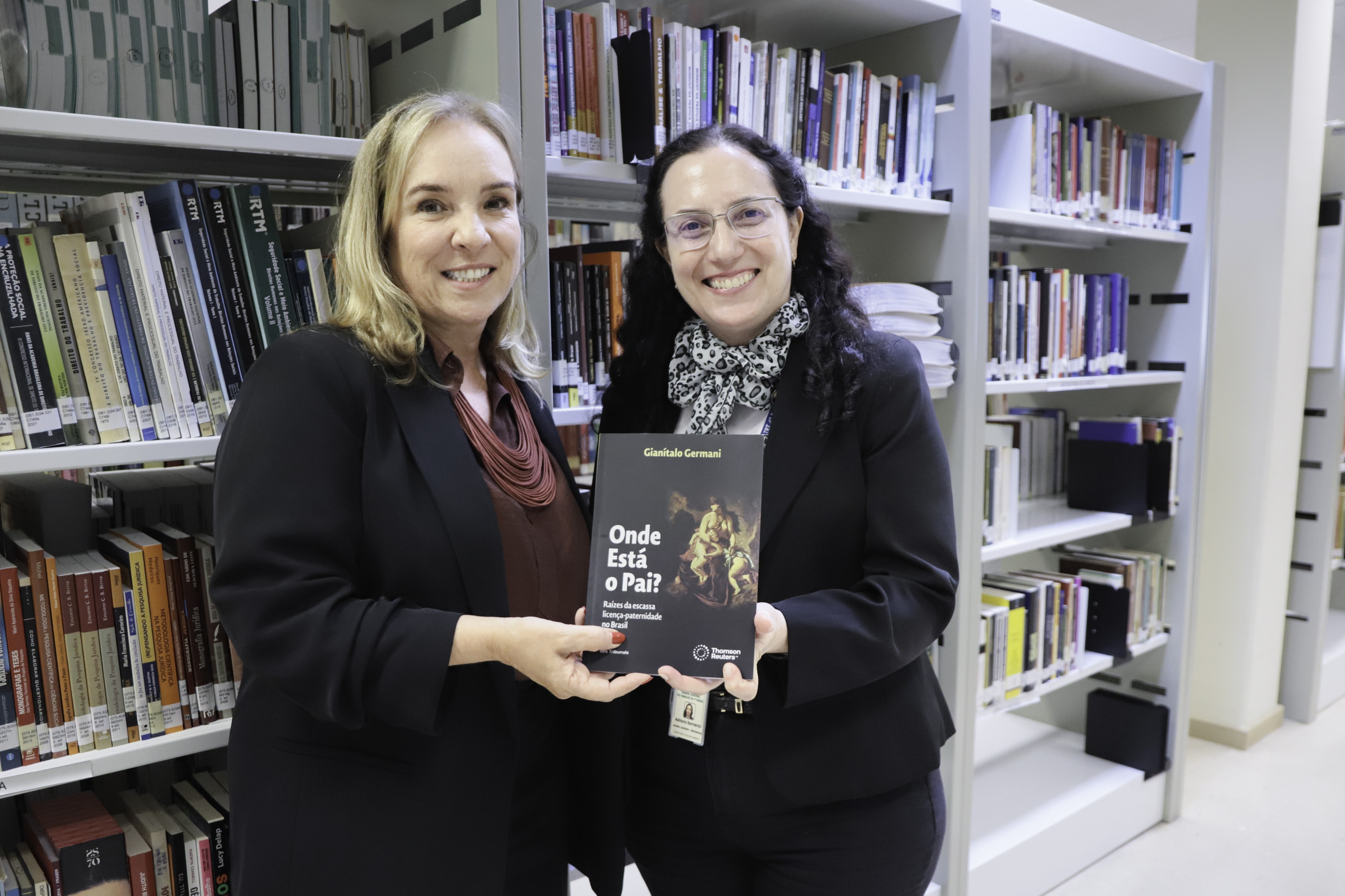 Maria Silvana e Adriana posam para a foto segurando um livro. Ao fundo aparece o acervo da biblioteca. 