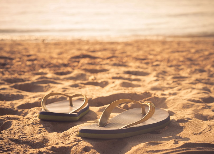Foto ilustrativa de um par de chinelos sobre areia.
