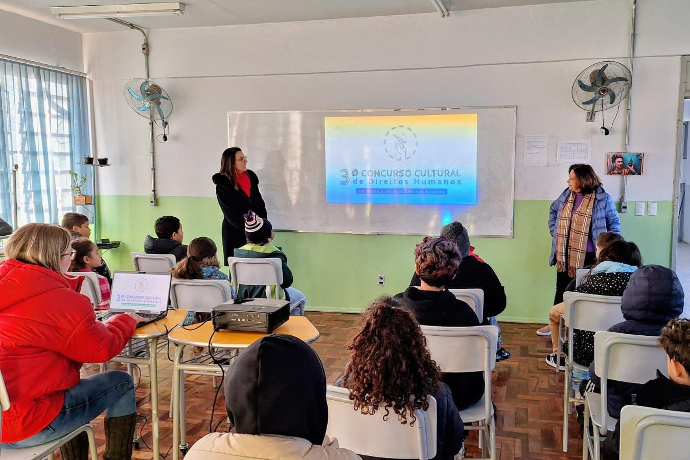 Sala de aula com alunos sentados de costas assistindo a uma apresentação. Ao fundo, no quadro, está projetada a pergunta: “Por que é importante combater o trabalho infantil?”. Márcio e Cassou falam com os estudantes; um deles está em pé ao centro e o outro próximo à porta. Sobre as mesas, há um notebook e um projetor.