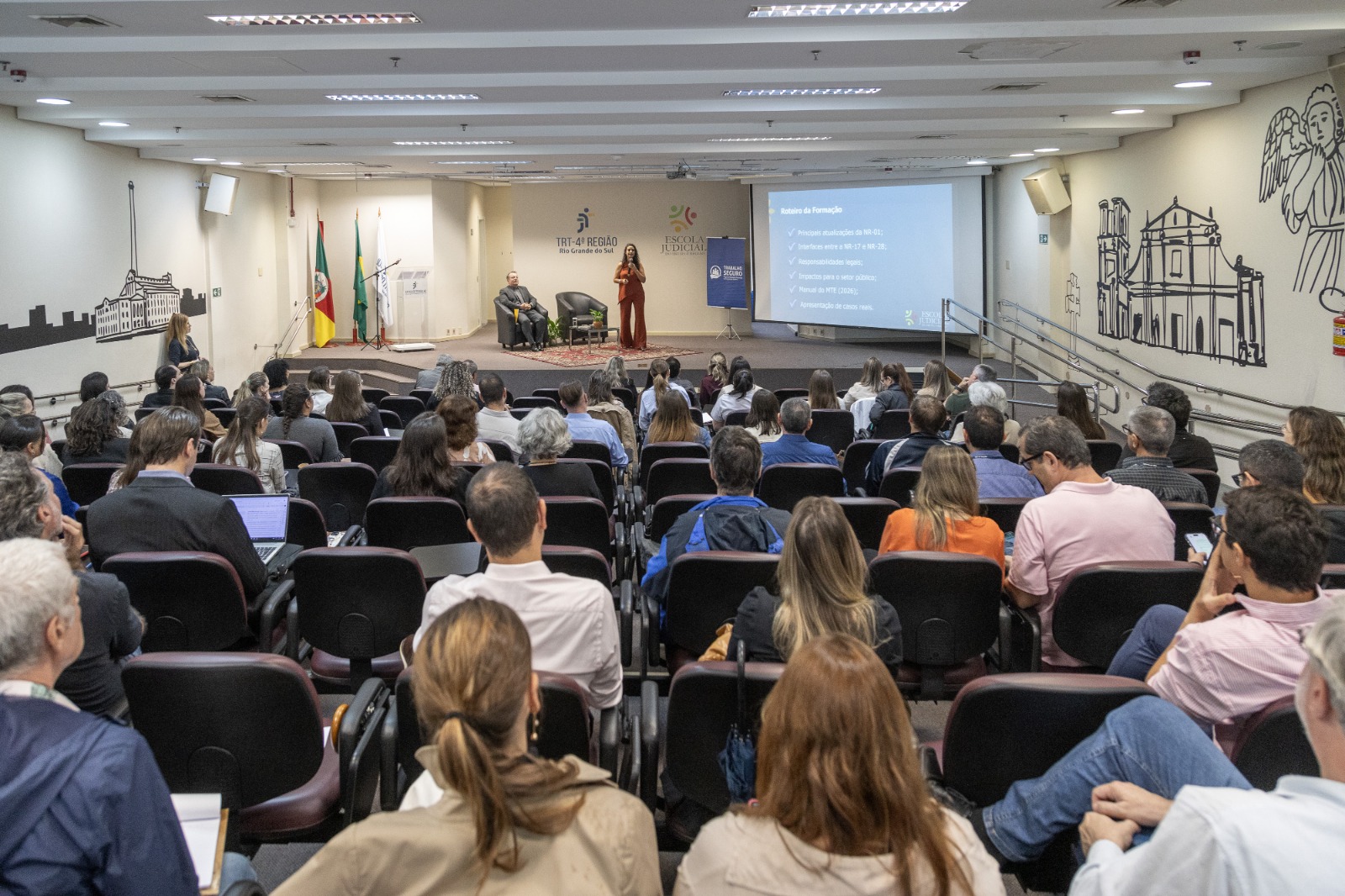 público em primeiro plano e palestrante ao fundo, falando de pé no palco