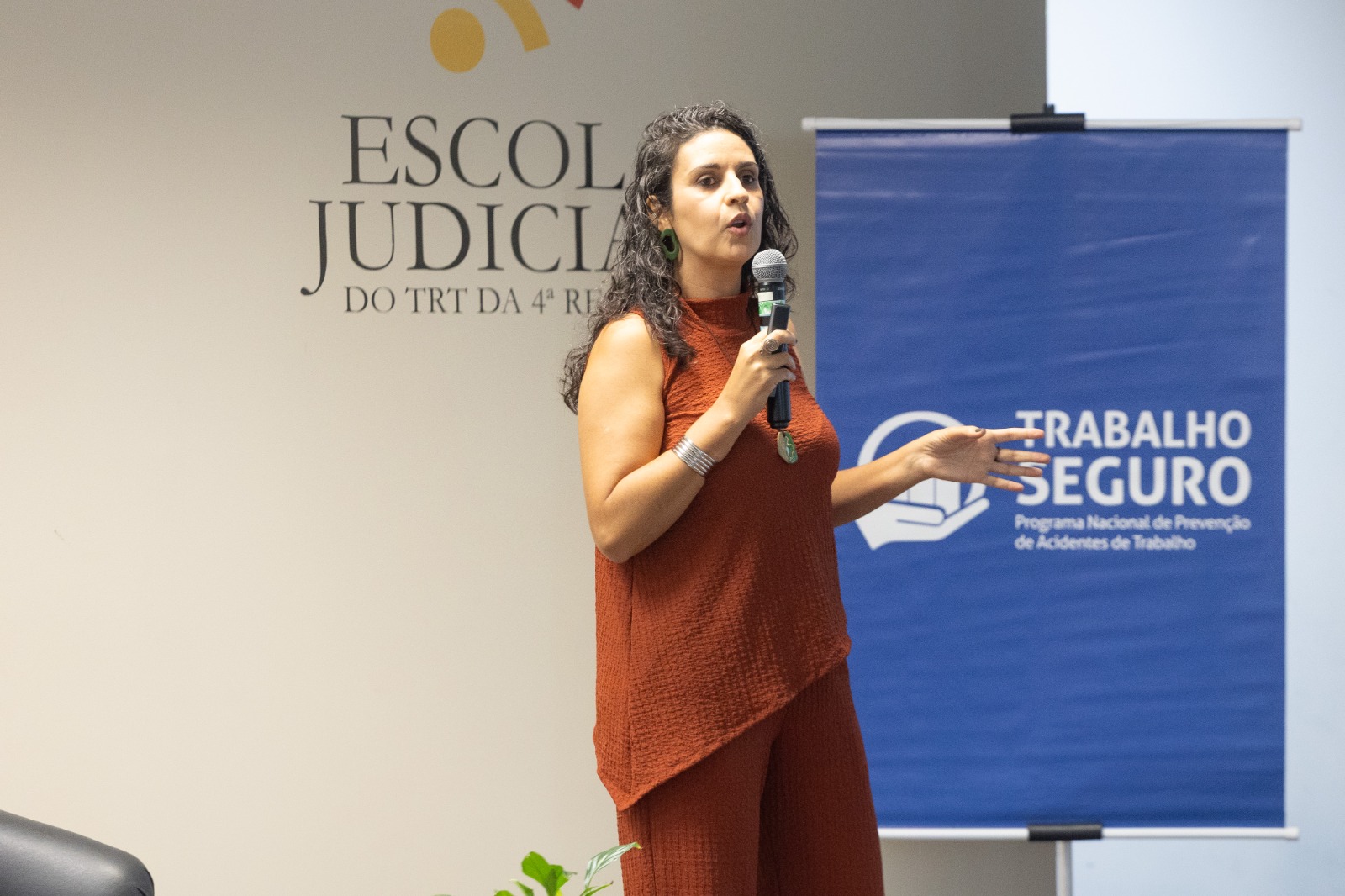 Kellen é parda, tem cabelo comprido castanho e usa blusa e calça laranja. fala de pé gesticulando e segurando um microfone.