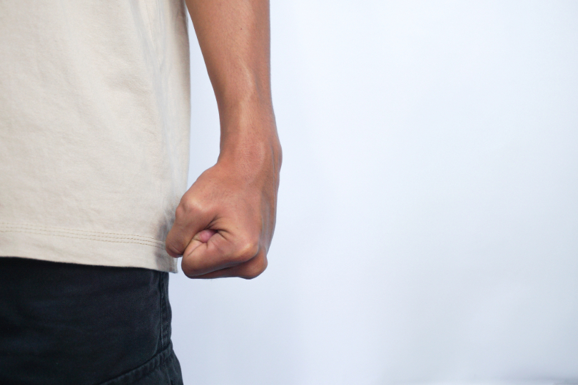 Mão masculina fechada, preparada para dar um soco. Apenas a mão do homem é mostrada, ao lado do corpo. Ele usa calça preta e camiseta branca. O fundo da imagem é branco.