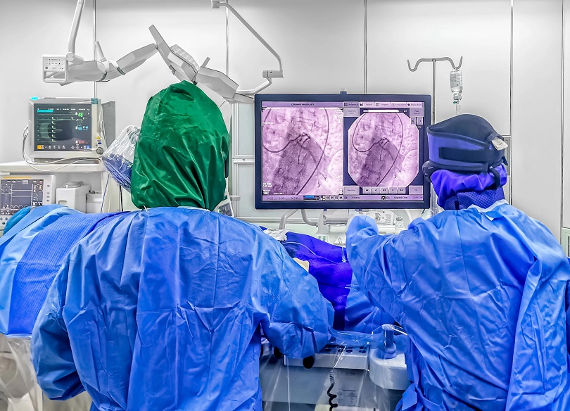 Foto ilustrativa mostra uma sala de cirurgia com dois profissionais de saúde realizando um procedimento médico. Eles estão de costas, usando roupas estéreis azuis, luvas e toucas, em um ambiente bem equipado.
