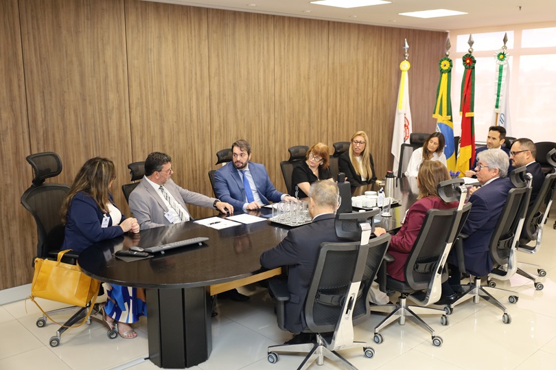 A fotografia mostra os participantes da reunião sentados aos redor de uma mesa oval e comprida, conversando.