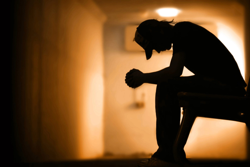 Silhueta de homem sentado, com a cabeça baixa, representando tristeza/ desespero. Predomínio de cor preta e amarelo escuro.