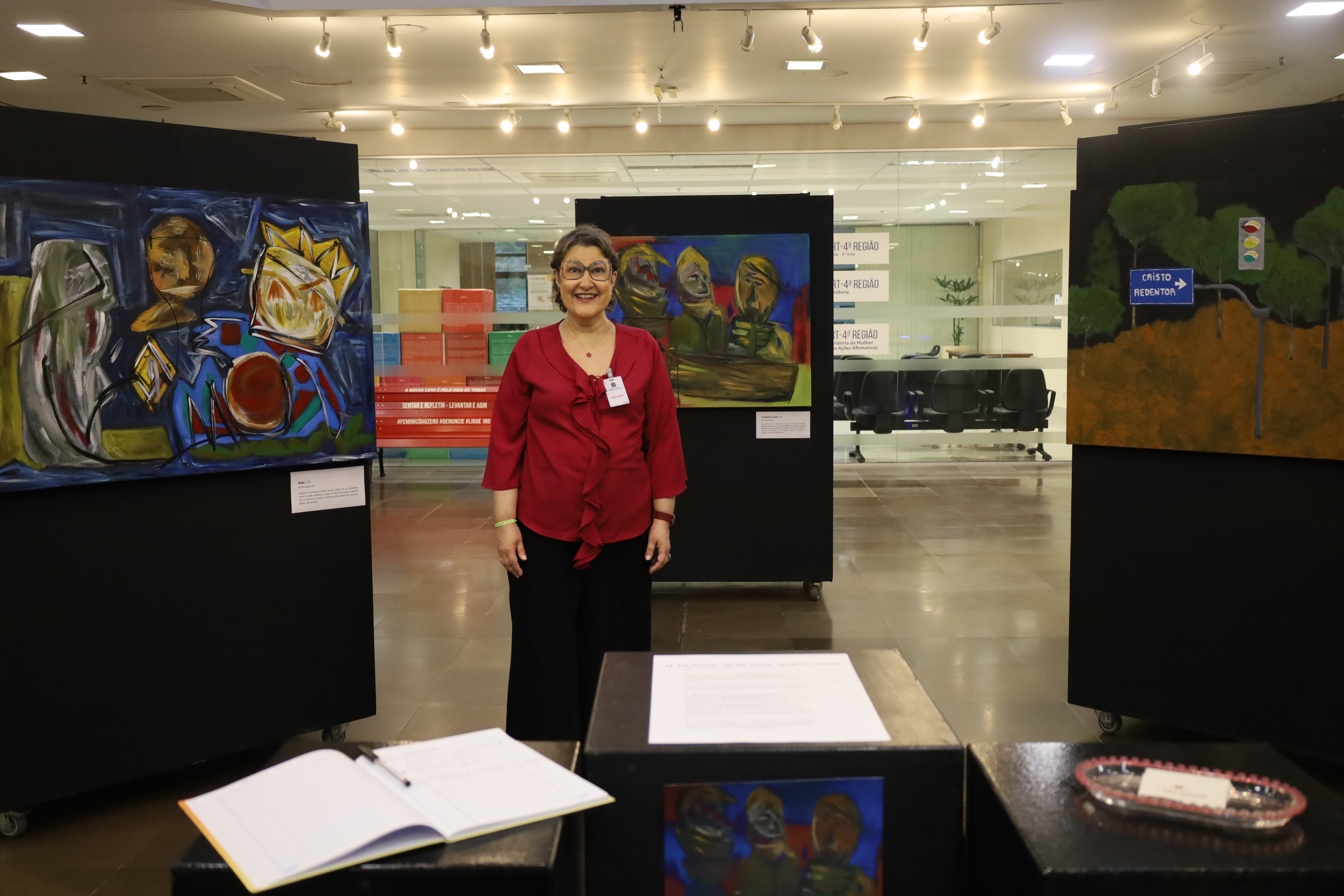 Silvia sorri no centro da exposição, vestindo uma blusa vermelha e calça preta. Atrás dela, três grandes telas aparecem em painéis pretos. Em primeiro plano, totens pretos exibem um livro de assinaturas aberto e materiais informativos.