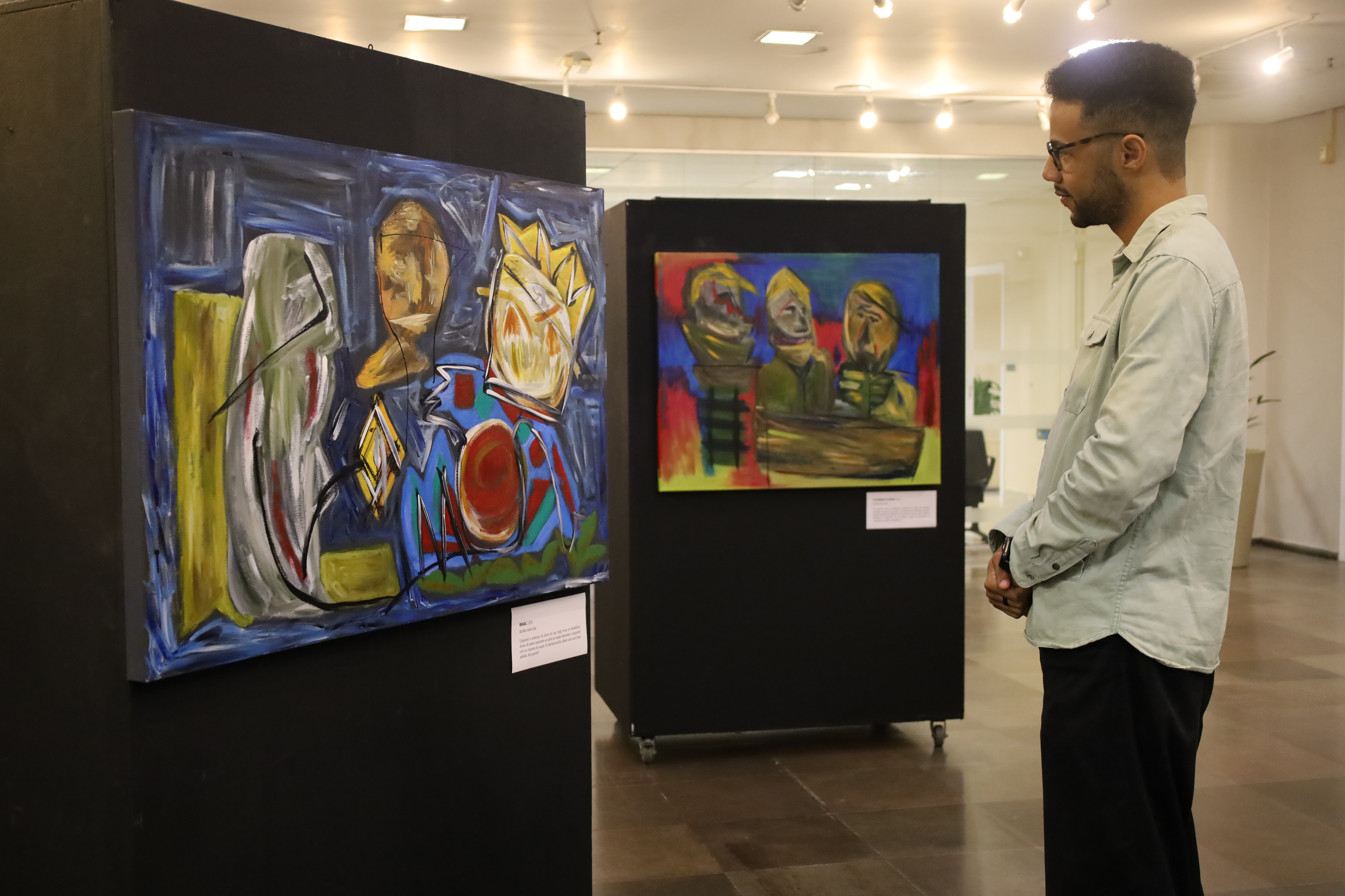 Um jovem de óculos e camisa clara observa atentamente, de perfil, a primeira obra da exposição. O foco recai sobre a pintura.