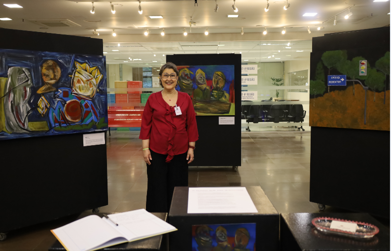 Silvia sorri no centro da exposição, vestindo uma blusa vermelha e calça preta. Atrás dela, três grandes telas aparecem em painéis pretos. Em primeiro plano, totens pretos exibem um livro de assinaturas aberto e materiais informativos.