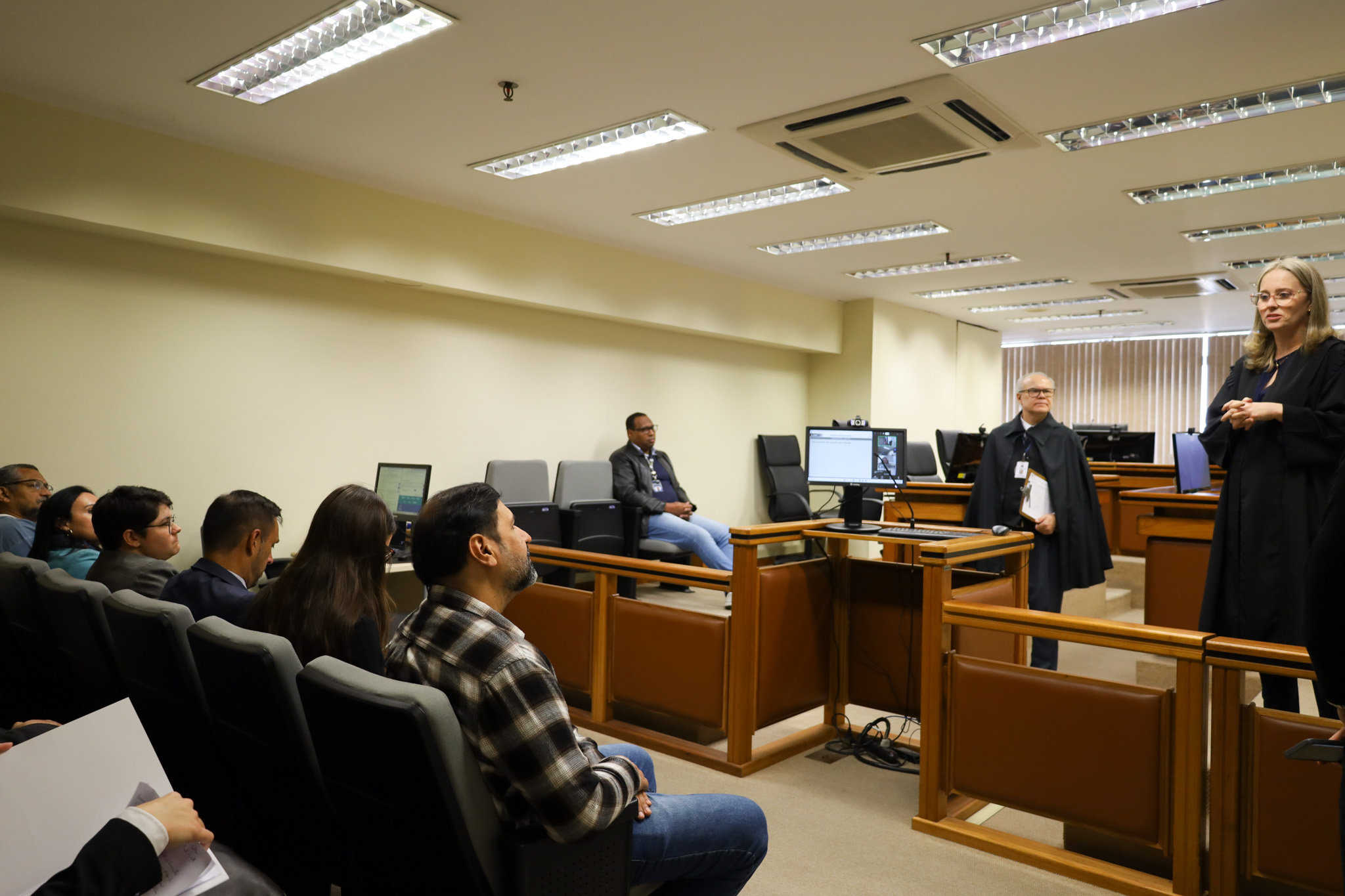 Fotografia lateral de uma sala de audiências. Em primeiro plano, estudantes estão sentados em poltronas cinzas, observando a cena. No centro, a secretária gesticula enquanto fala ao grupo. Próximo a ela, um homem também de toga segura uma prancheta.