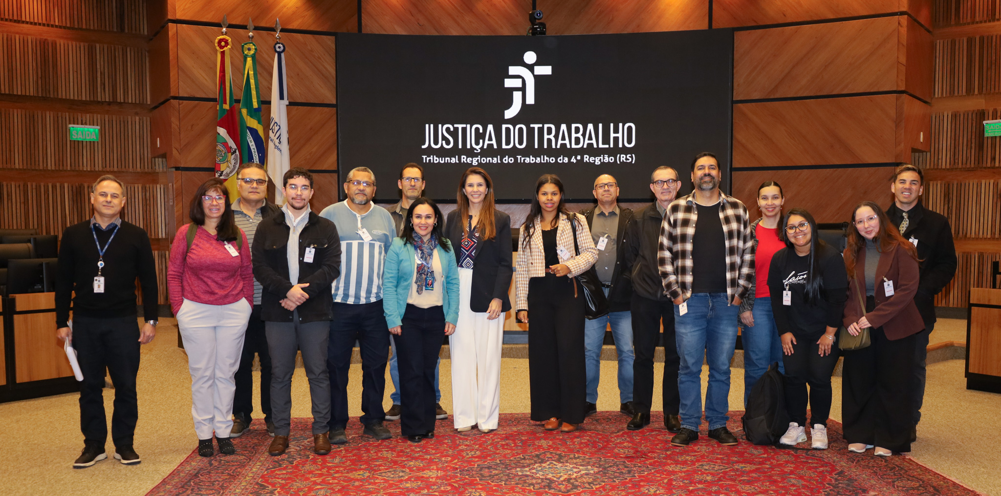 Duas alunas posam para a foto, uma veste um blazer xadrez e carrega uma bolsa preta, a outra usa um casaco marrom. Elas estão ao lado do secretário de audiência e de um magistrado. 