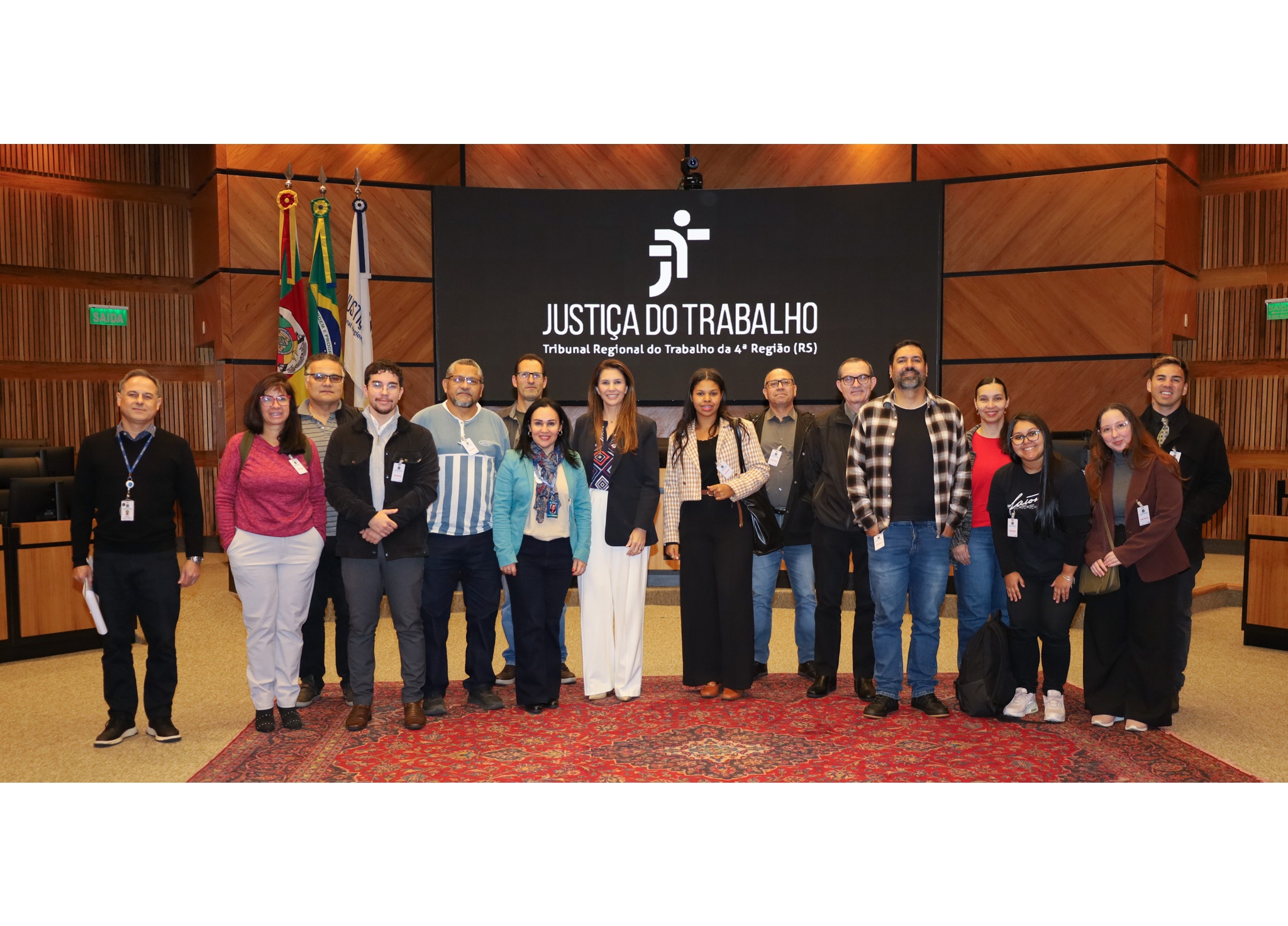 O grupo de 15 pessoas posam para uma foto no plenário, eles estão posicionados sobre um tapete avermelhado.