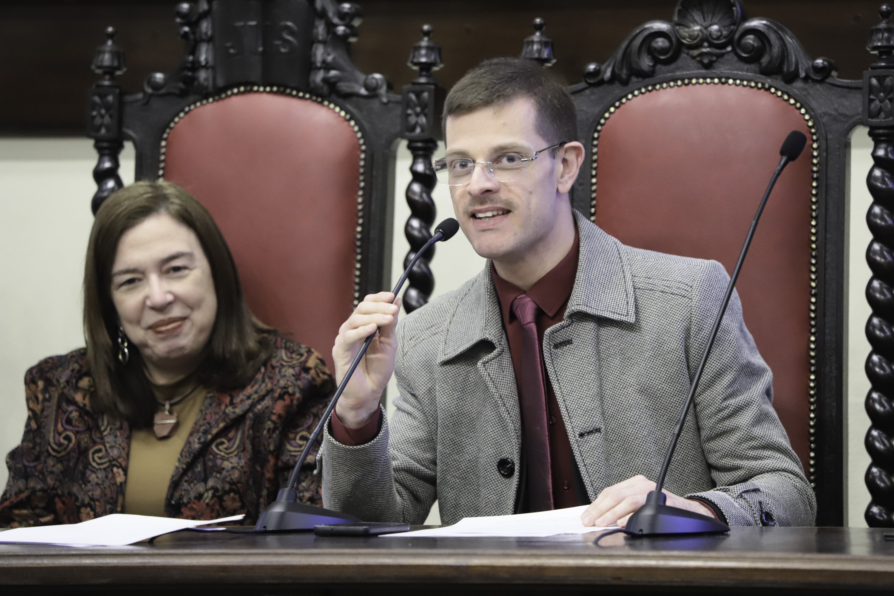 Ana Paula sorri na esquerda e Guilherme fala na direta ao microfone. Ambos estão na mesa oficial