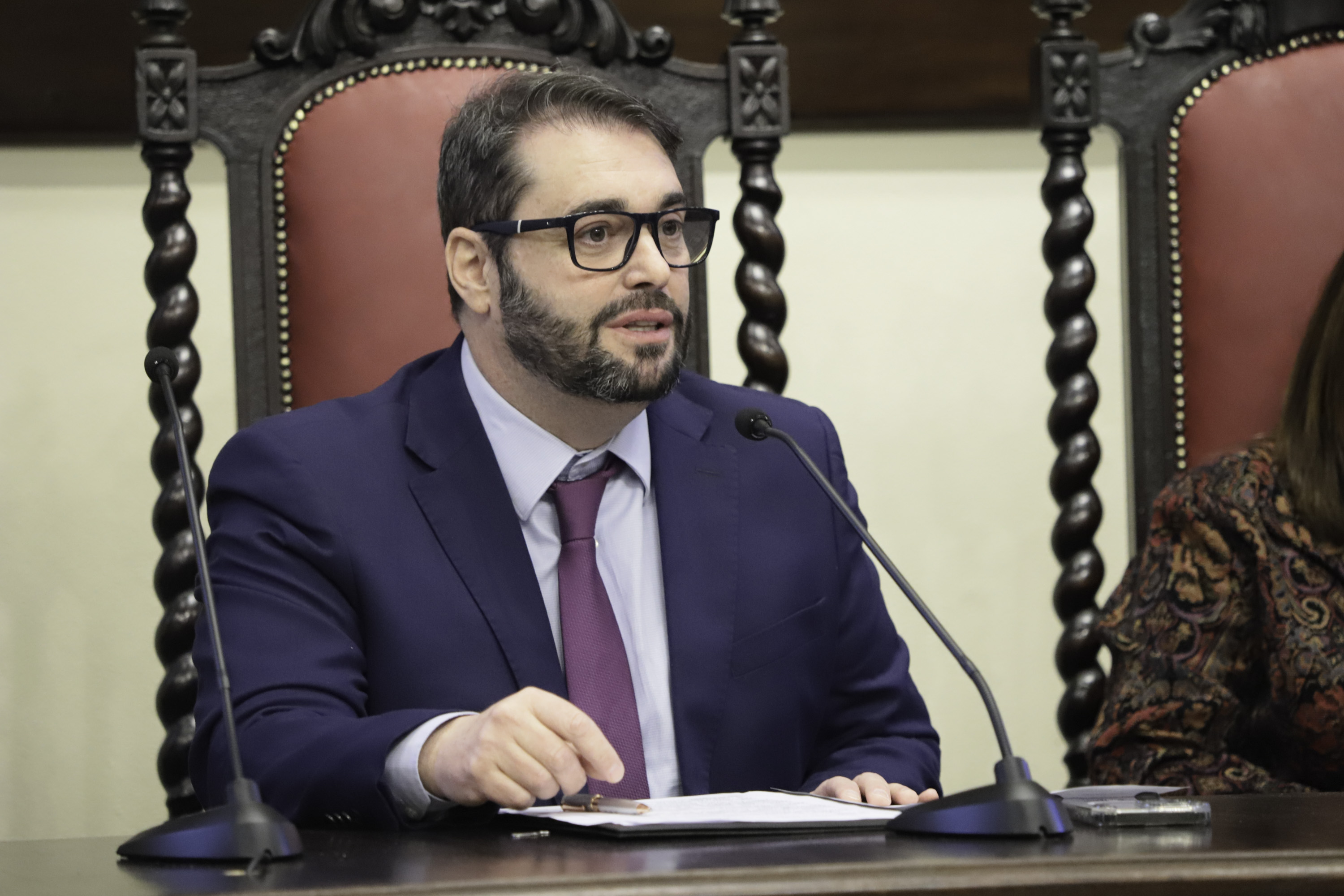 Alexandre é branco, usa cabelo e barba escura, óculos de aro preto e fala na mesa oficial. 