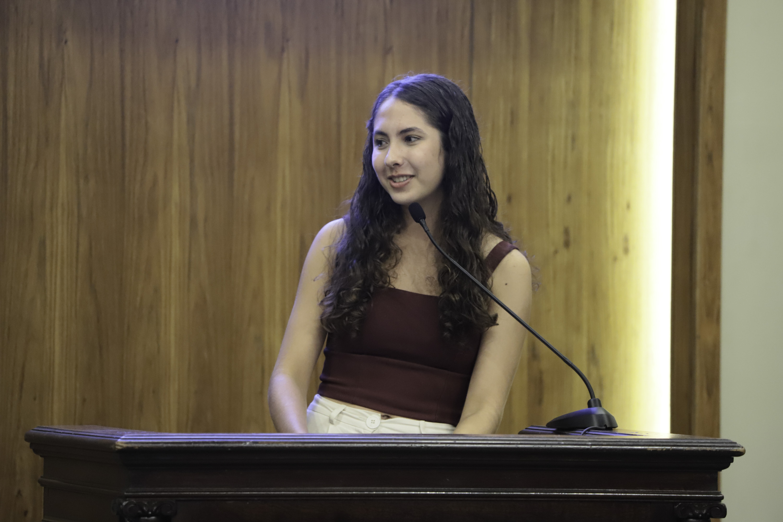 Manuela é parda de cabelo comprido escuro, fala no púlpito olhando para a esquerda. 