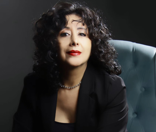 Alejandra é uma mulher de cerca de 50 anos, de pele clara e cabelos pretos e crespos. Ela usa batom vermelho e blusa e terno pretos.