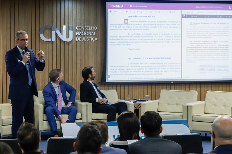 Frederico está de pé, na esquerda da imagem, falando e gesticulando ao microfone. Na direita, aparece um telão com o sistema Galileu ao fundo e o público de costas em primeiro plano.