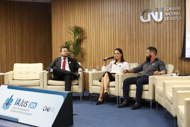 Natacha aparece sentada em um sofá falando ao microfone. 