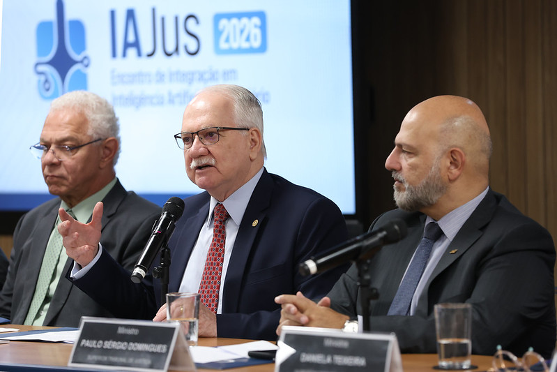 Fachn fala e gesticula ao microfone, no centro da imagem. 