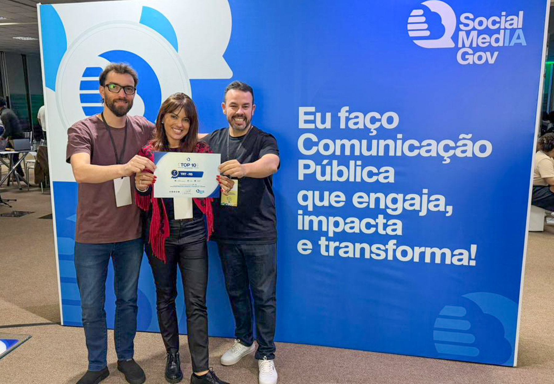 Daniel Aguiar, Camila Dotto e Eduardo Matos, com o certificado na mão, em frente ao banner do evento.