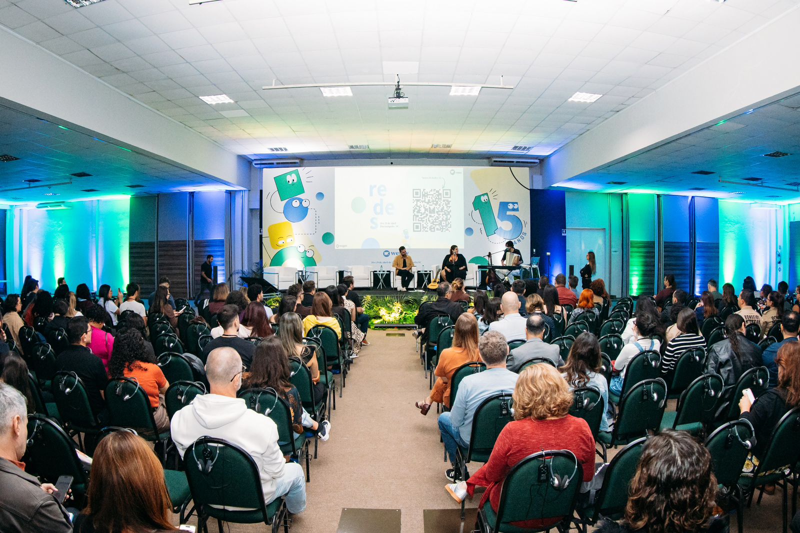 Foto em angulo aberto, mostrando palestrantes no palco e público assistindo