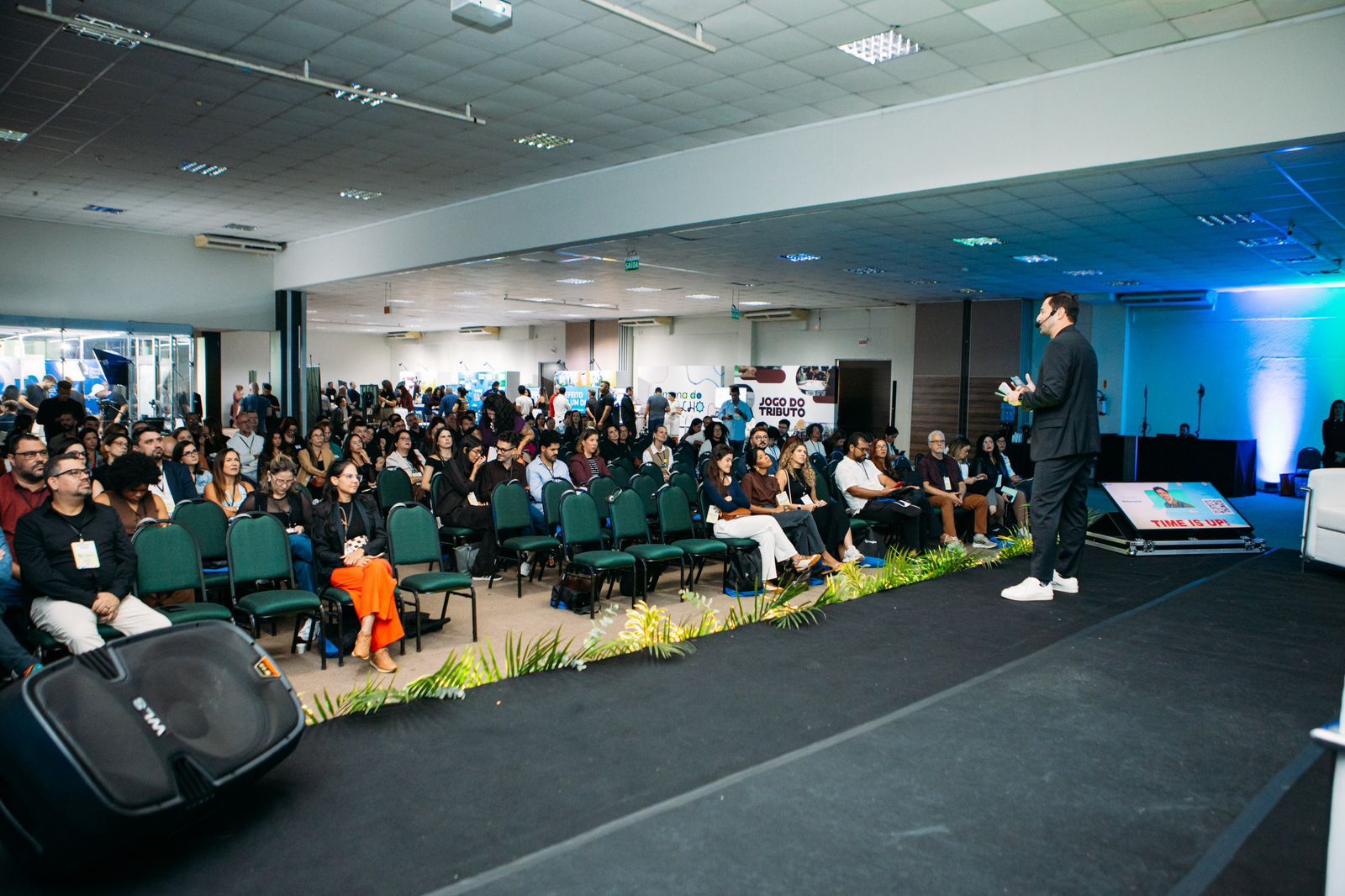 Foto em angulo aberto, mostrando palestrantes no palco e público assistindo