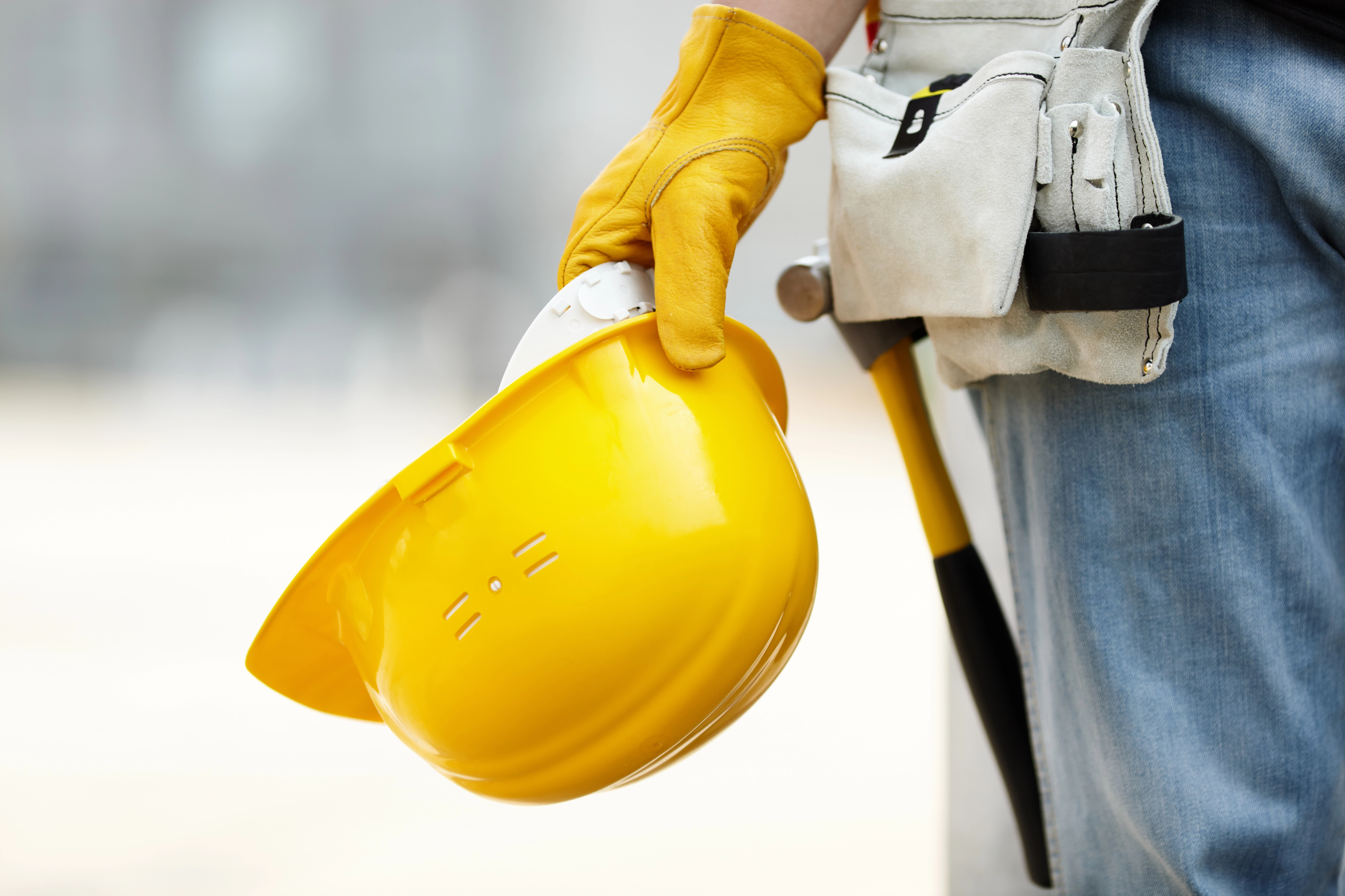 Em primeiro plano, uma mão protegida por uma luva de couro amarela robusta segura firmemente um capacete de segurança amarelo brilhante pela aba. O trabalhador veste calças jeans azuis e utiliza um cinto de ferramentas de camurça bege na cintura, onde se nota o cabo de um martelo amarelo e preto.