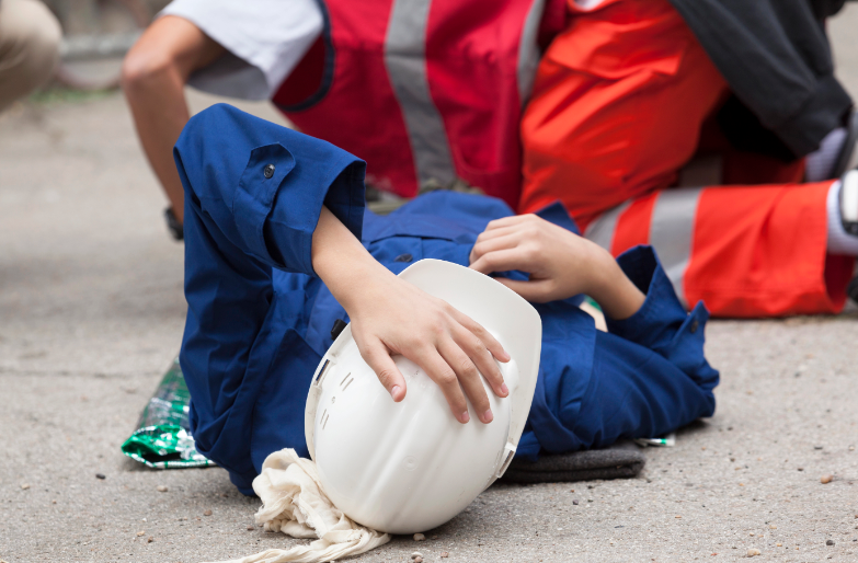 Uma pessoa vestindo um macacão de trabalho azul-escuro está deitada de costas sobre o asfalto cinza, com o braço erguido segurando um capacete branco sobre o rosto. Ao fundo, um socorrista em trajes de alta visibilidade (vermelho e laranja) está ajoelhado para prestar assistência. Uma manta térmica verde metalizada aparece dobrada ao lado do corpo.