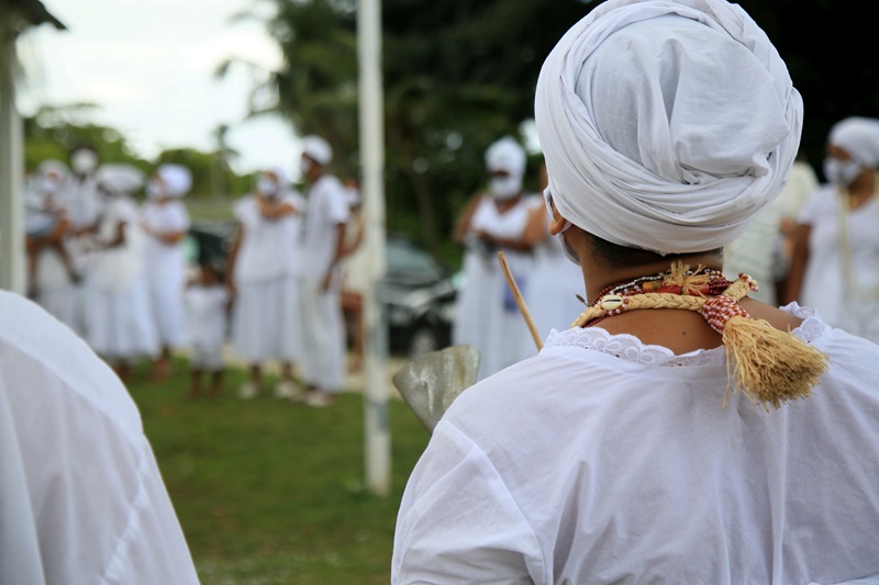 Fotografia em close em uma pessoa, de costas, vestindo roupas brancas de uma religião de matriz africana.