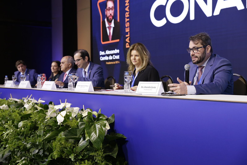 Foto em primeiro plano do presidente palestrando, com demais painelistas ao lado, na mesa do painel.
