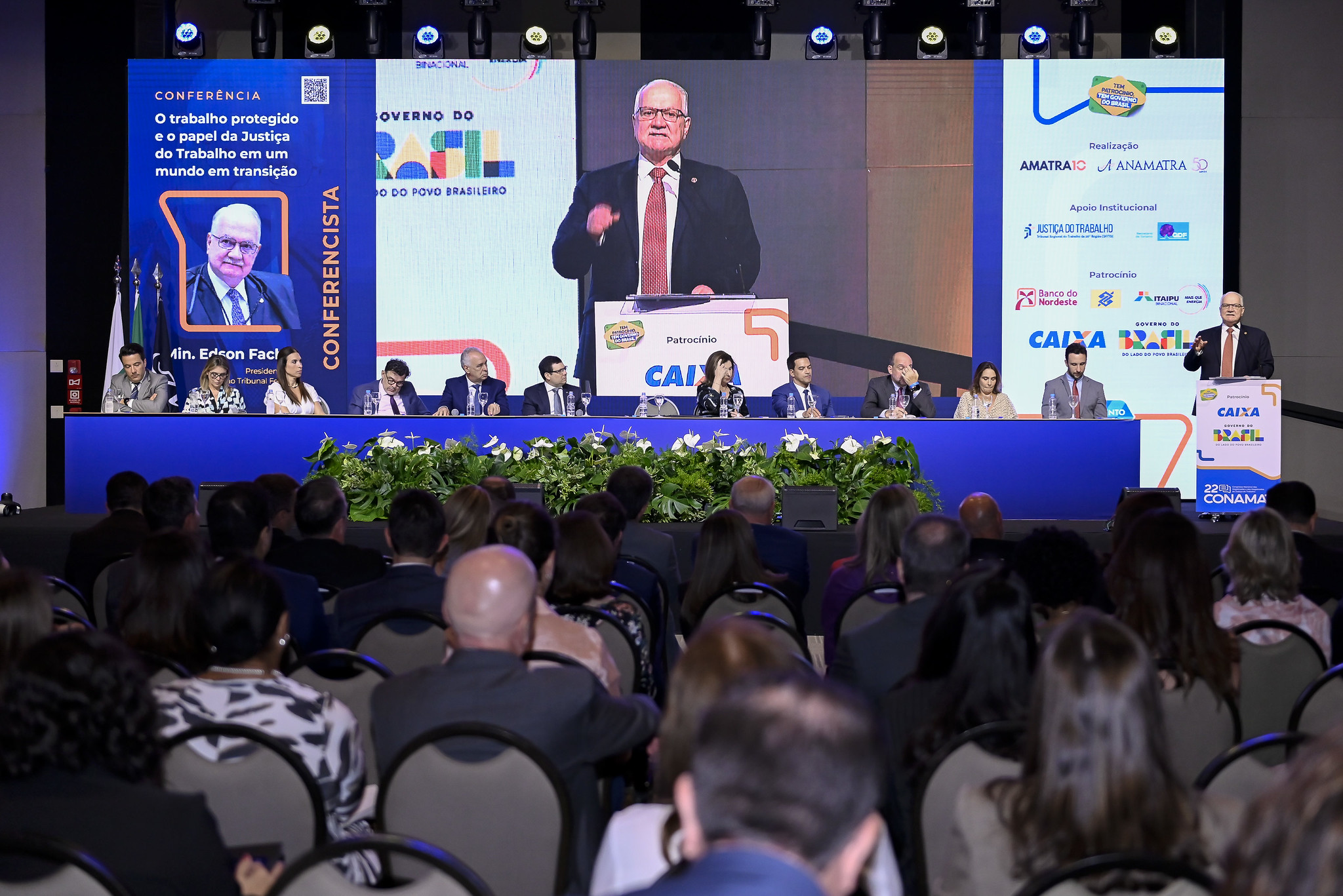 A foto mostra o ministro Fachin num telão e várias pessoas no plano inicial na frente.