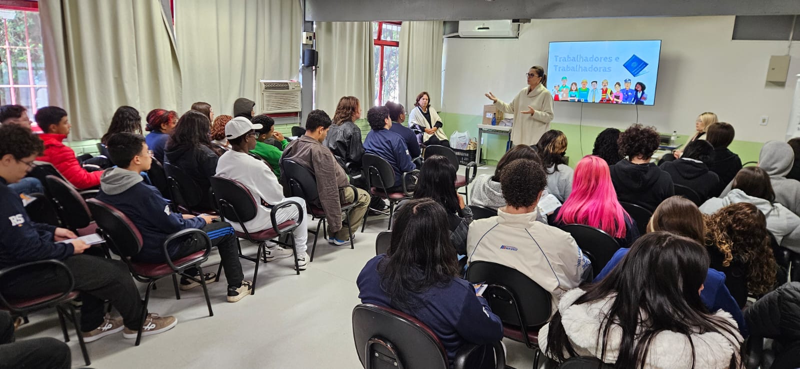 Juíza Aline gesticulando enquanto fala para uma plateia de estudantes. Ao fundo, uma televisão exibe um slide azul com o título "Trabalhadores e Trabalhadoras", ilustrado com diversos personagens representando profissões. Os adolescentes estão sentados em cadeiras pretas, muitos de costas para a câmera, demonstrando atenção.