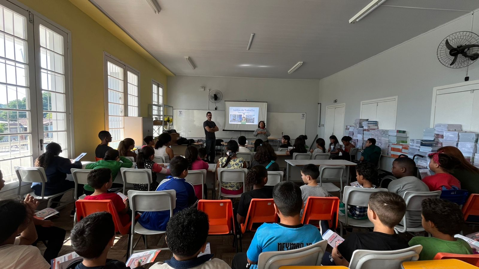 A imagem mostra uma sala de aula ampla e bem iluminada por grandes janelas laterais. À frente, Rejane e Rafael conduzem uma atividade. O projetor exibe o tema "Justiça do Trabalho". A plateia é composta por crianças e pré-adolescentes sentados em classes escolares com cadeiras brancas e laranjas. No lado direito, veem-se pilhas de livros didáticos organizadas.