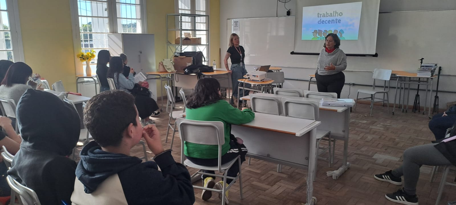 Em uma sala com piso de taco de madeira e grandes janelas, Rejane fala com alunos. O slide projetado ao fundo destaca o termo "trabalho decente" com figuras coloridas. Laís auxilia próximo ao projetor.