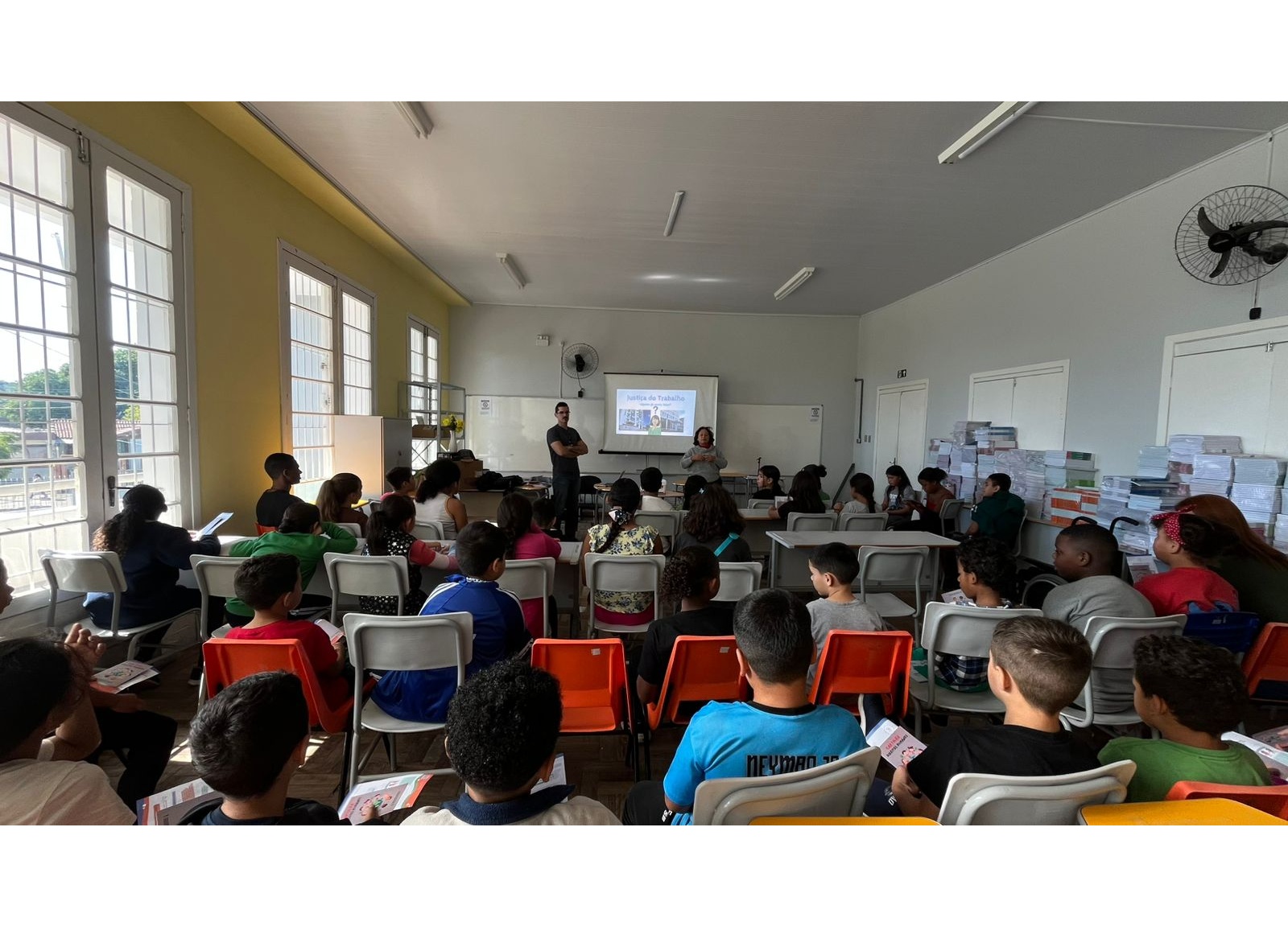 A imagem mostra uma sala de aula ampla e bem iluminada por grandes janelas laterais. À frente, Rejane e Rafael conduzem uma atividade. O projetor exibe o tema "Justiça do Trabalho". A plateia é composta por crianças e pré-adolescentes sentados em classes escolares com cadeiras brancas e laranjas. No lado direito, veem-se pilhas de livros didáticos organizadas.