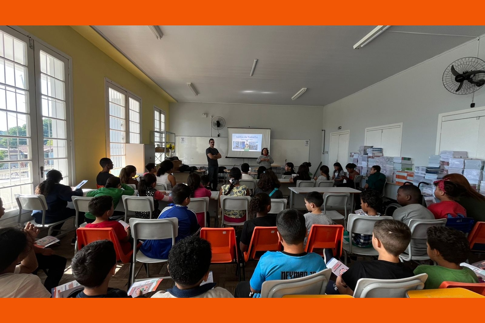 A imagem mostra uma sala de aula ampla e bem iluminada por grandes janelas laterais. À frente, Rejane e Rafael conduzem uma atividade. O projetor exibe o tema "Justiça do Trabalho". A plateia é composta por crianças e pré-adolescentes sentados em classes escolares com cadeiras brancas e laranjas. No lado direito, veem-se pilhas de livros didáticos organizadas.