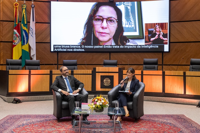 A fotografia mostra a palestrante Gabrielle Sarlet, que tem sua imagem projetada em um telão, e sentados estão o palestrante João Sérgio e a juíza Luciana Xavier.
