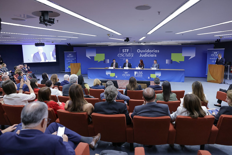 A fotografia mostra a plateia do evento, sentada, de costas, e mais ao longe a mesa de abertura, composta pelo ministro Edson Fachin