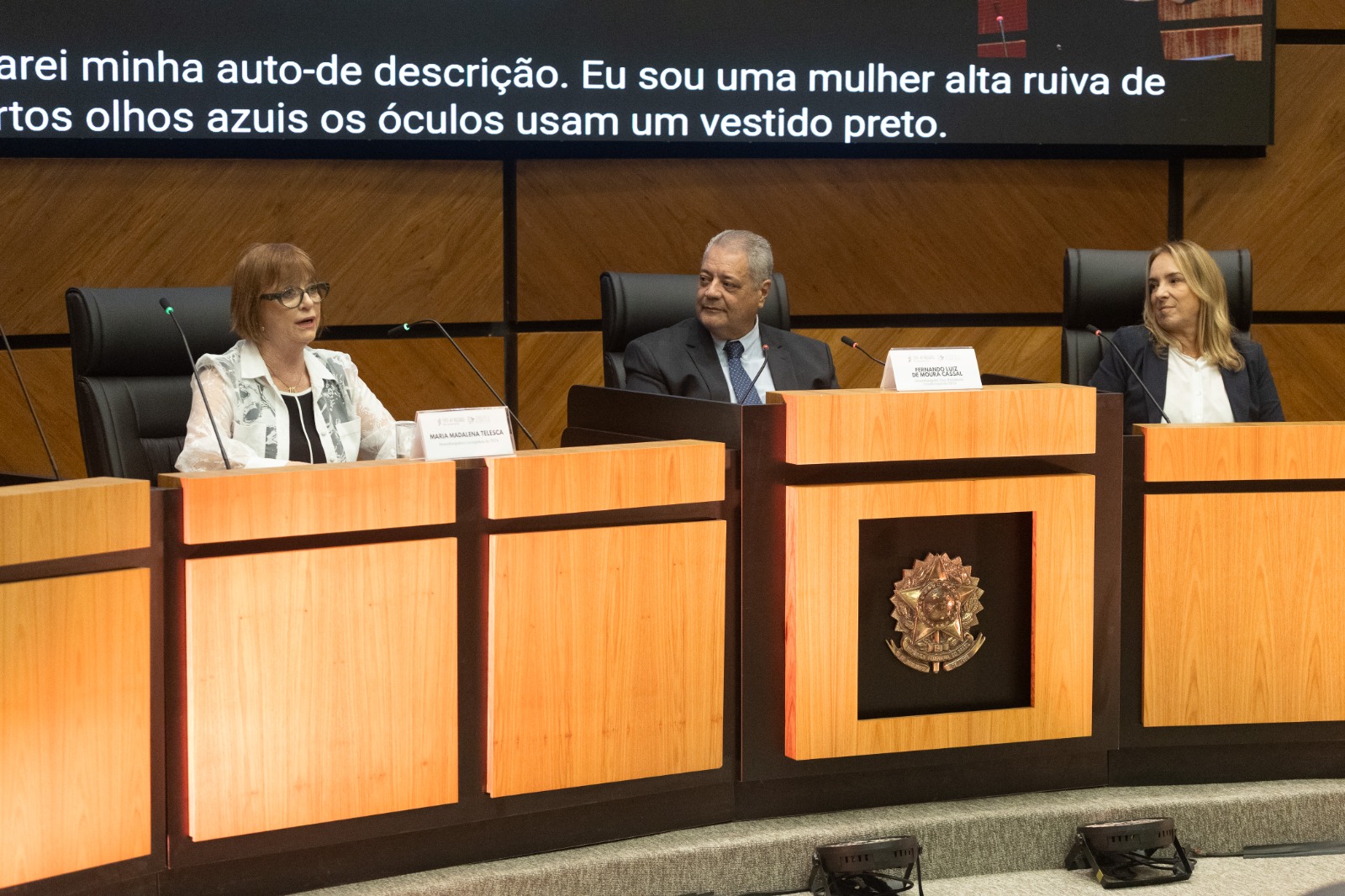 três pessoas na mesa oficial do evento com telão ligado ao fundo