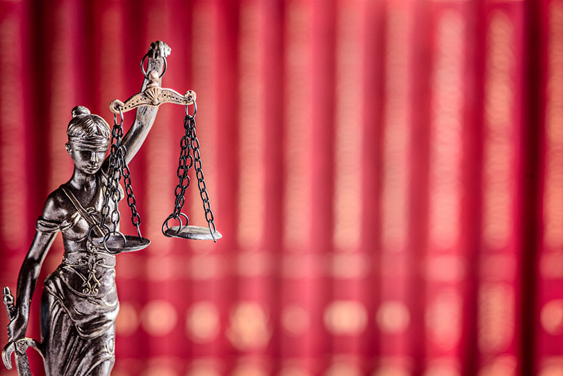 Pra Cego Ver: foto de djedzura/iStock banco de imagens mostra pequena estátua da deusa da Justiça, Themis. Ao fundo estão livros na cor vermelha.