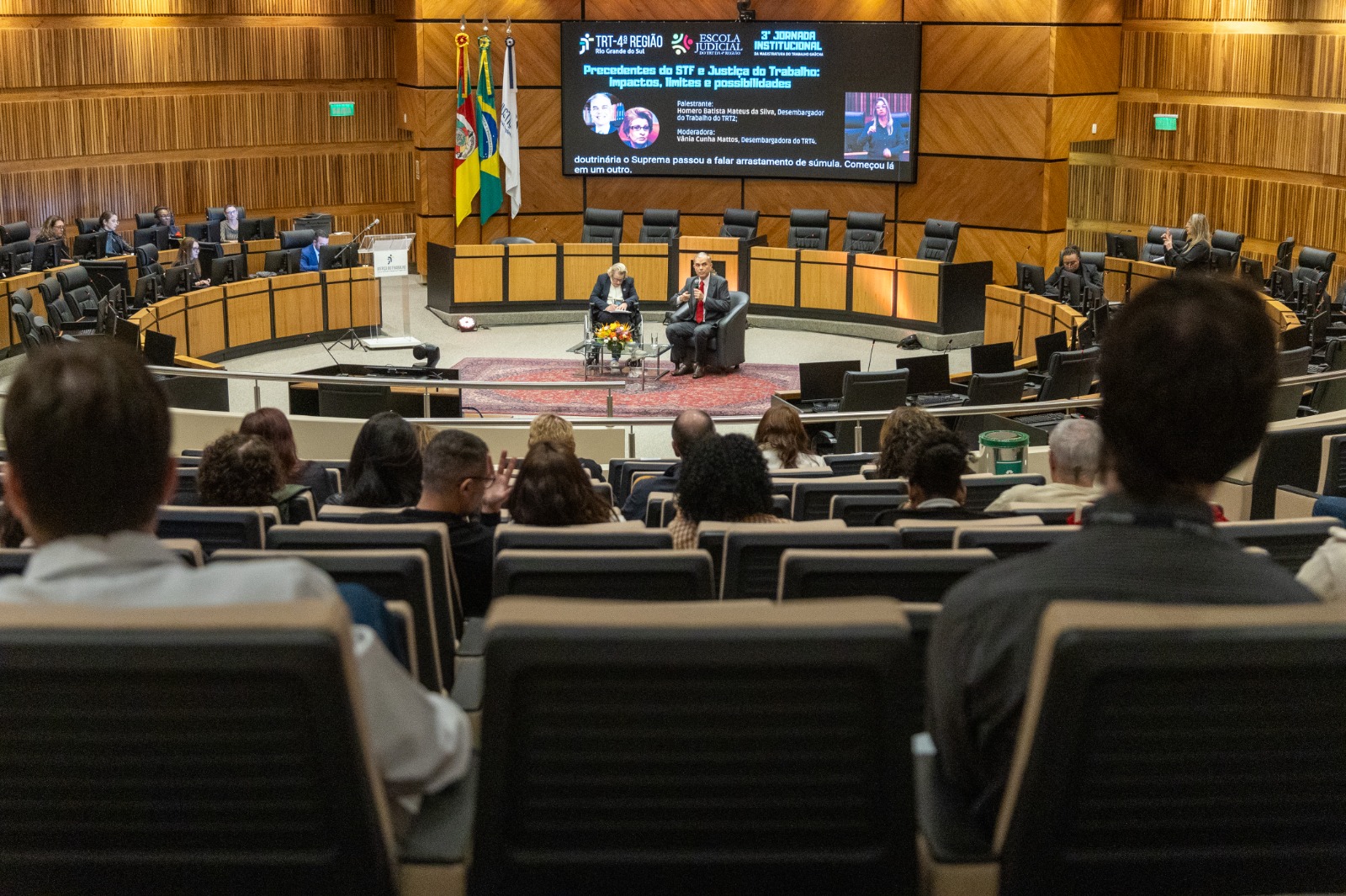 Foto geral do plenário com público de costas e dois palestrantes ao fundo